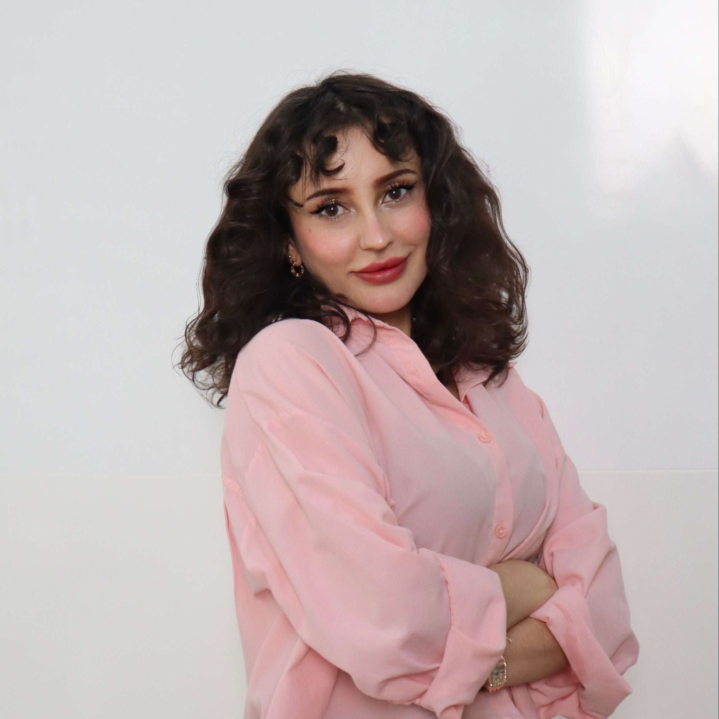 Smiling Şeyda Çevik with shoulder-length curly dark hair, wearing a light pink blouse, posing with her arms crossed against a white background.