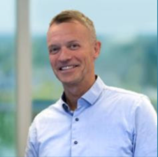 Smiling Svein Erik Nicolaysen, wearing a light blue button-up shirt, standing indoors with large windows showing a blurred outdoor view in the background.
