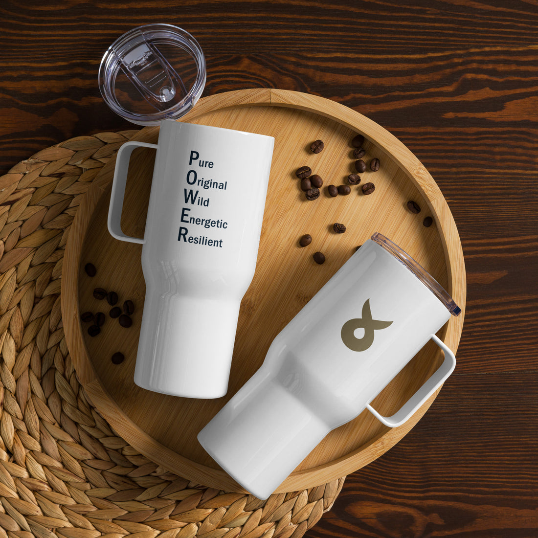 Two white travel mugs with handles and transparent lids placed on a wooden tray surrounded by coffee beans. One mug features the acronym "POWER" with the words "Pure, Original, Wild, Energetic, Resilient" printed vertically. The other mug displays a gold Ballstad logo.